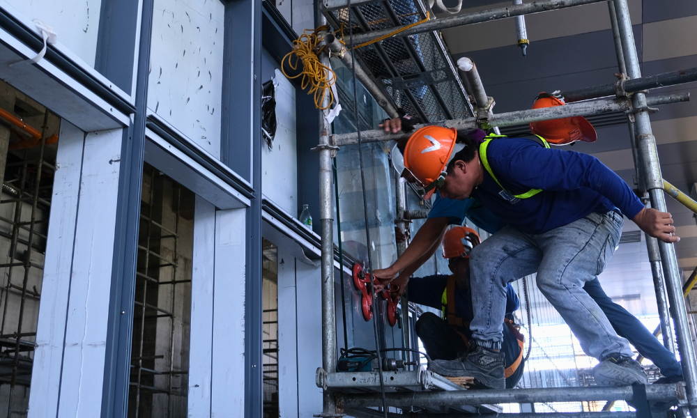 A construction worker in a blue shirt and full safety gear, standing on scaffolding. In a partially constructed building