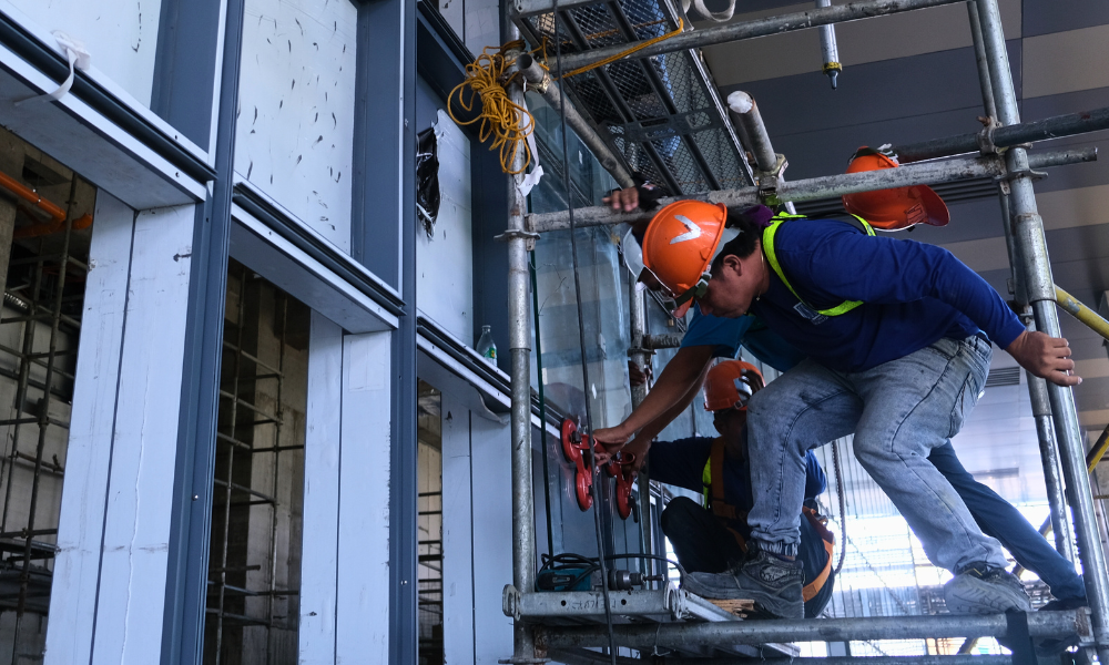 A construction worker in a blue shirt and full safety gear, standing on scaffolding. In a partially constructed building 